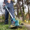 Werkzeuge – Qualität für Profis Eine Person, die mit einem akkubetriebenen Rasentrimmer Gras und Unkraut in einem Gartenbereich mit Bäumen und Blumen in der Nähe mäht. Jetzt kaufen