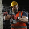 Werkzeuge – Qualität für Profis Ein Mann mit gelbem Schutzhelm und orangefarbener Sicherheitsweste bearbeitet mit einer Akku-Bohrmaschine eine Betonwand in einer Industrieumgebung. Jetzt kaufen